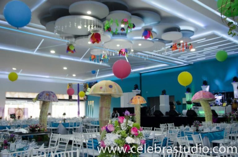 Salón de eventos decorado con globos de colores y setas gigantes sobre las mesas vestidas de blanco